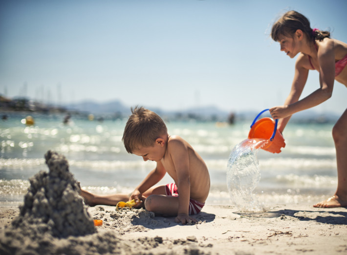 Little boy is playing on a beach while his sister has sneaked behind hims with a bucket of water and is just pouring the water on him.