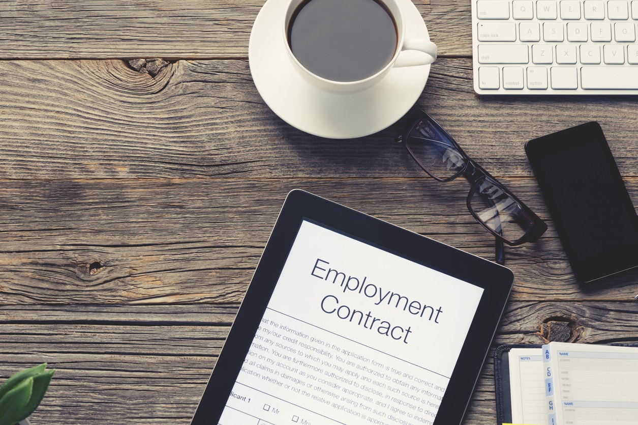 Online employment or job contract. The document is displayed on a digital tablet. There is also a mobile phone, computer keyboard, coffee, Filofax organizer notepad and glasses on the table. The table is made of wood and is quite old. Copy space on left.