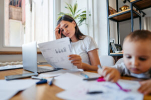 Tired Mid Adult Woman Working On The Laptop While Her Son Is Drawing On The Dining Table